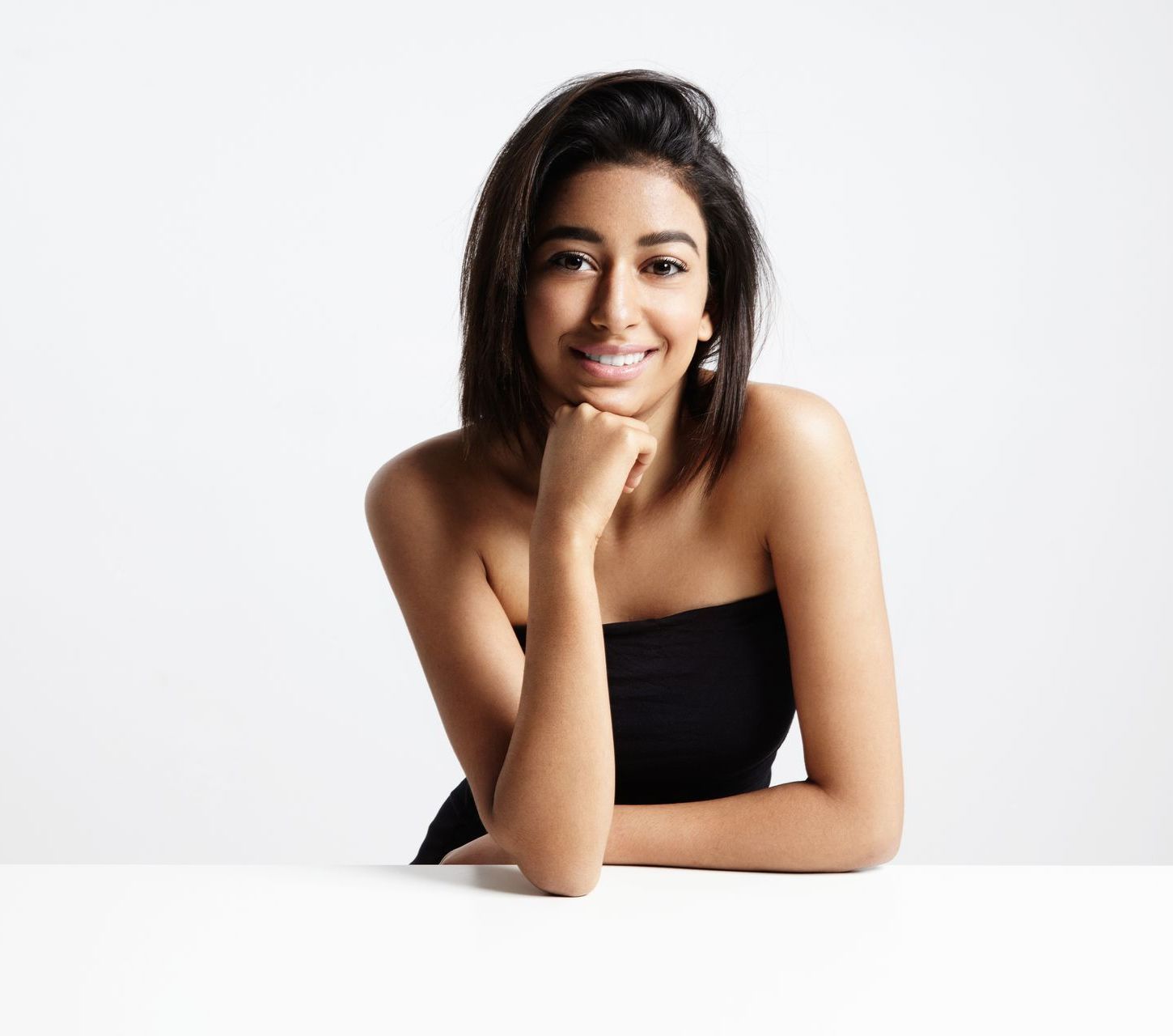 A woman is sitting at a table with her hand on her chin and smiling.