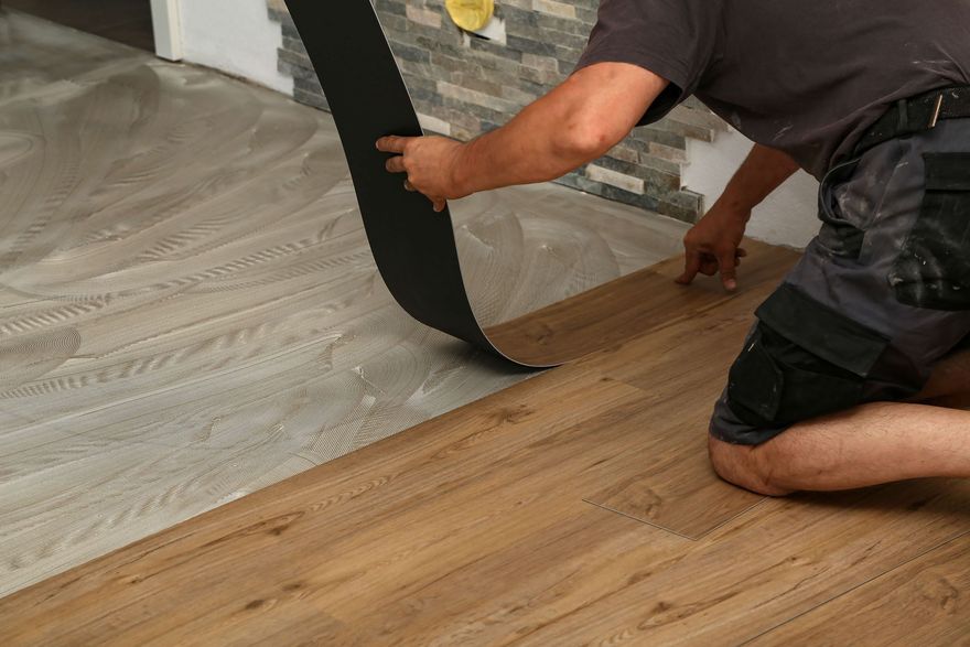 Worker installing wood flooring beside gray tile floor, fitting a plank in place.