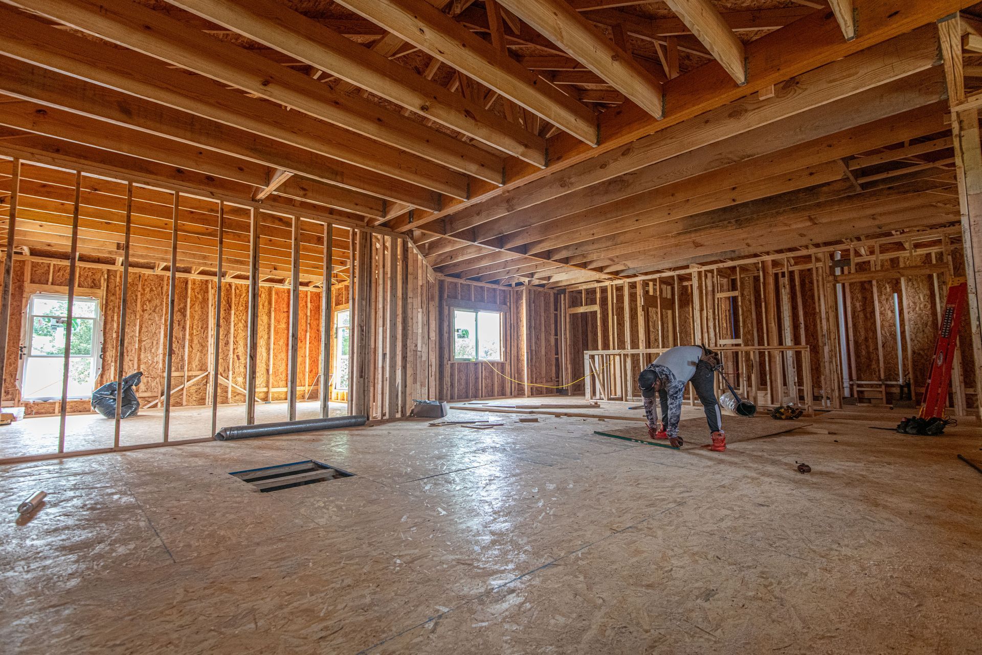 Interior of a framed house under construction, with exposed beams and a worker bending over the floor.
