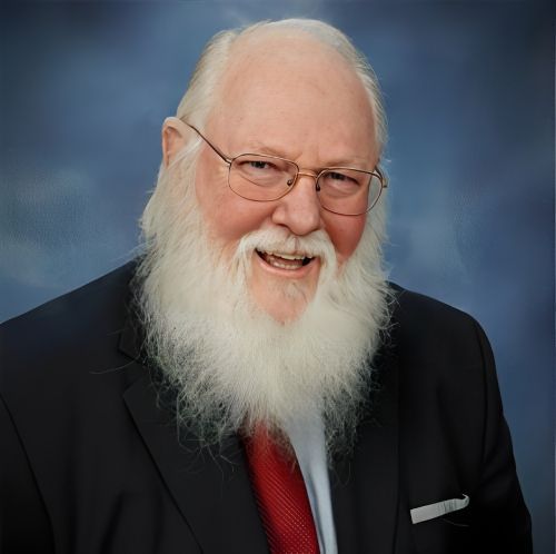 Man with a long white beard and glasses, wearing a suit and red tie, smiles at the camera against a blue background.