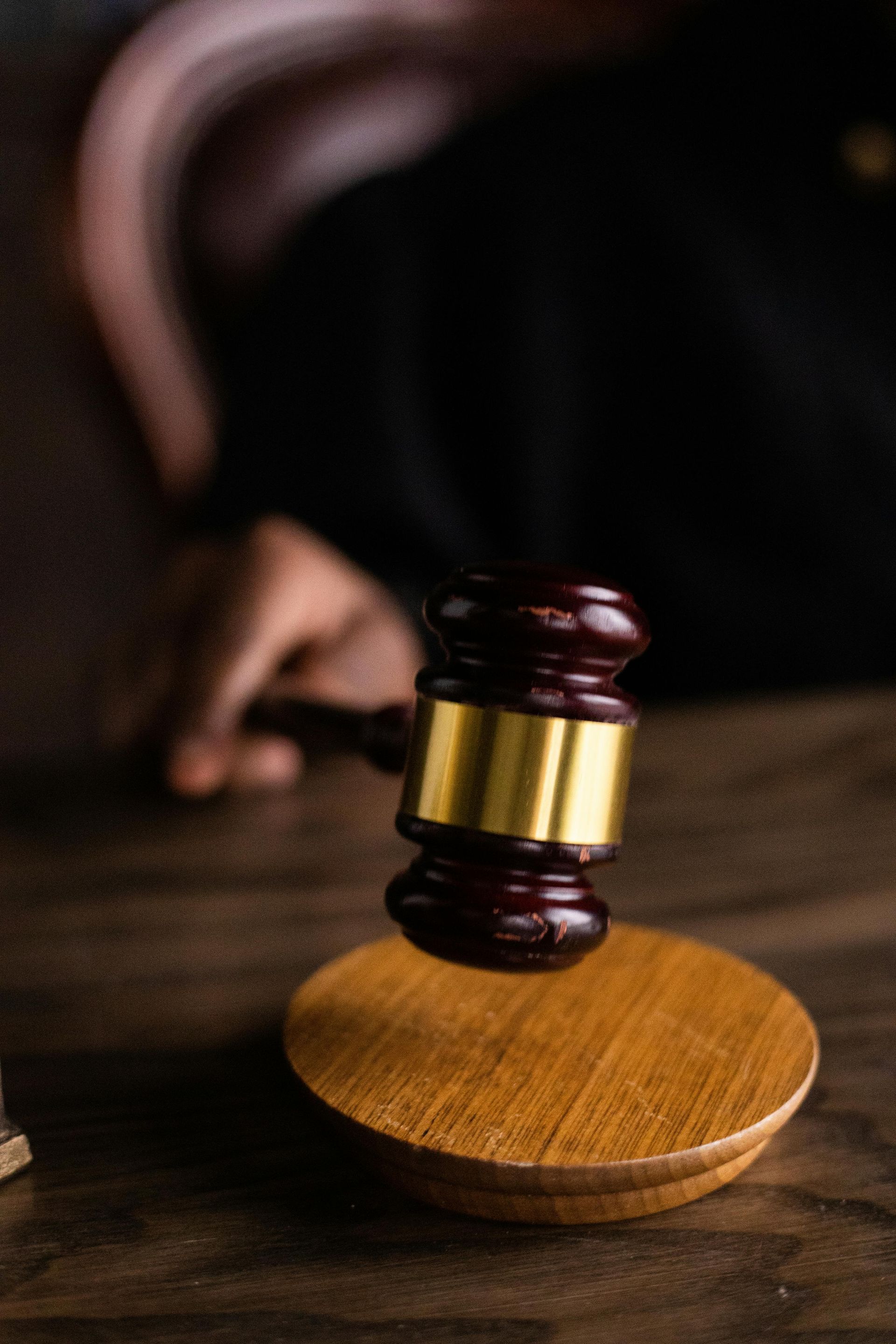 A judge's gavel on a wooden block, poised above a dark table. A person in a black robe is partially visible.