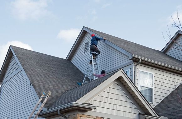 Two people on ladders working on a house roof. One person is on the roof, the other is near the ground.