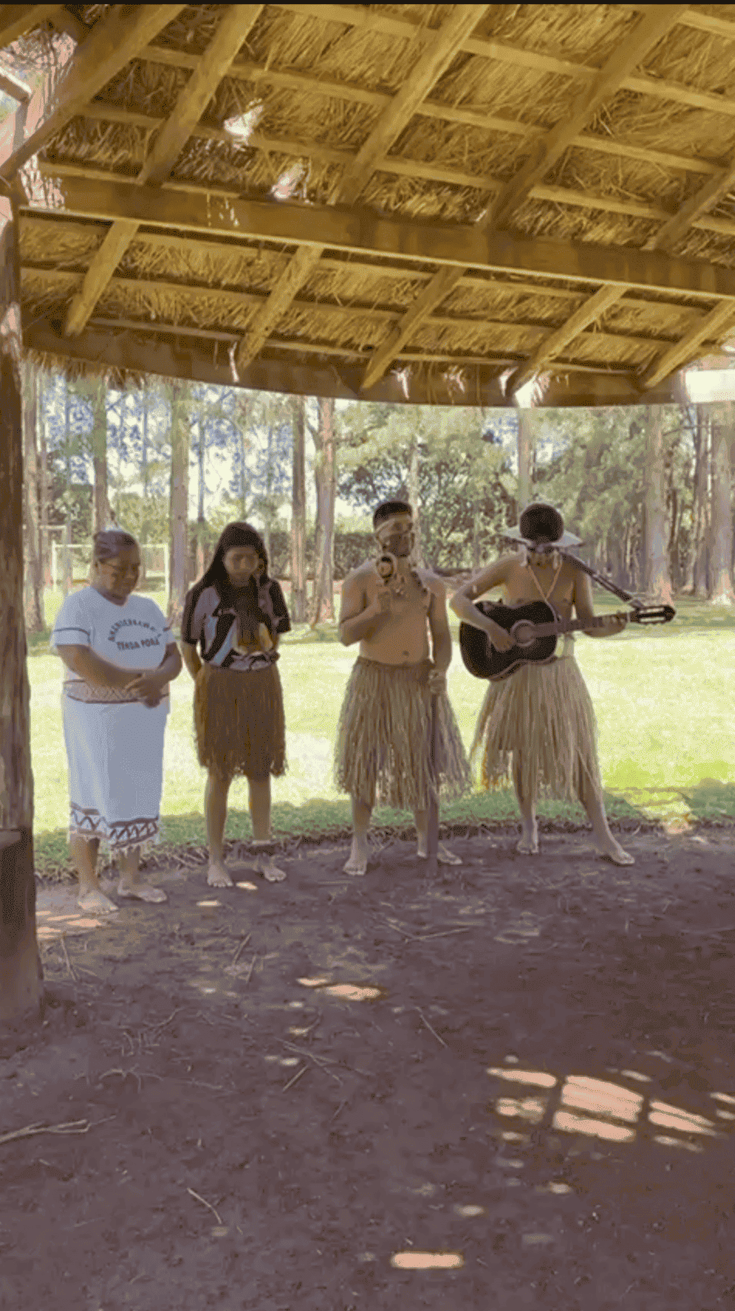 Um grupo de pessoas está cantando e tocando violão sob um teto de palha.