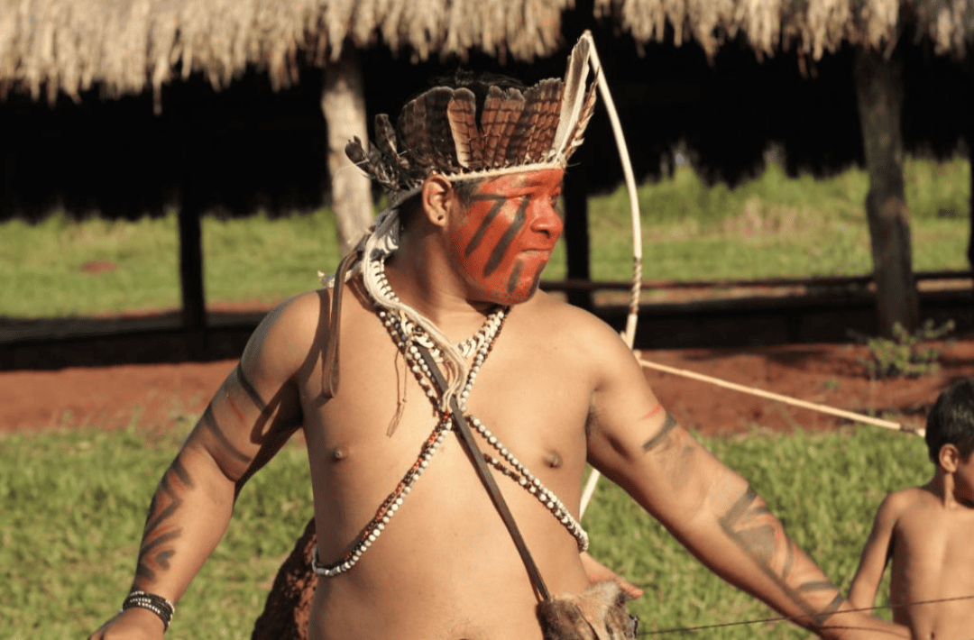 Um homem sem camisa com tinta vermelha no rosto está parado em um campo