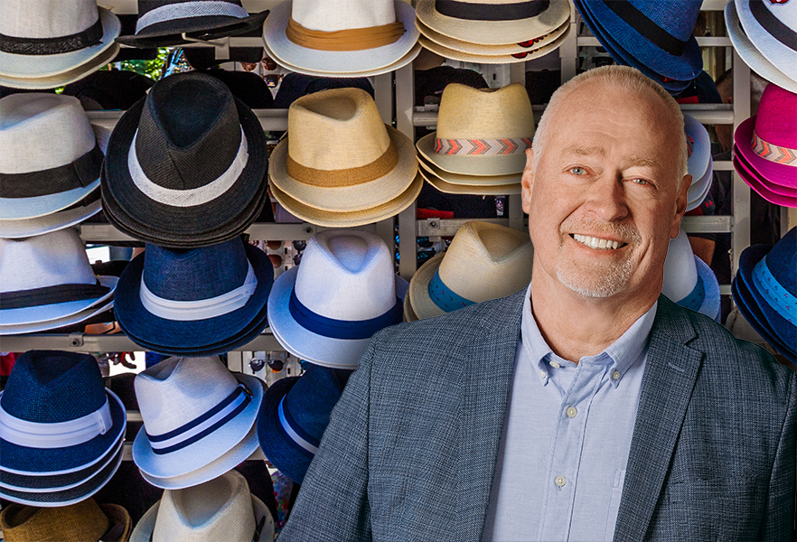 Mand smiler foran en væg af fedorahatte; han har en blå jakke og skjorte på.