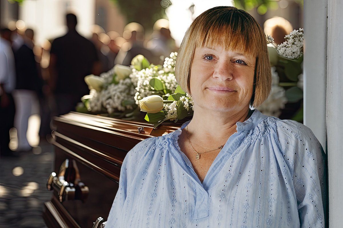 Kvinde i blå skjorte ved siden af ​​en kiste med hvide blomster; mennesker i baggrunden.