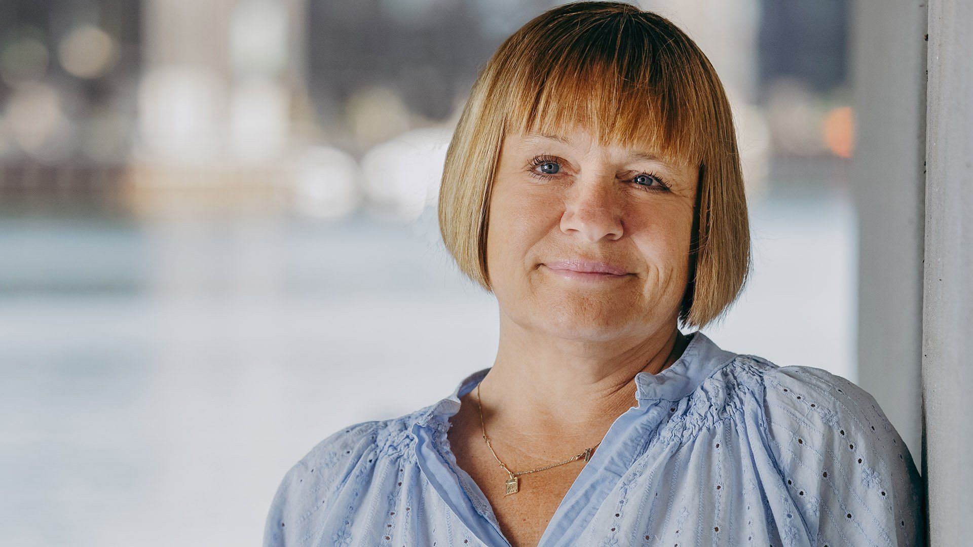 Kvinde i blå skjorte, lænet op ad en hvid væg, smilende. Sløret baggrund af vand og bygninger.