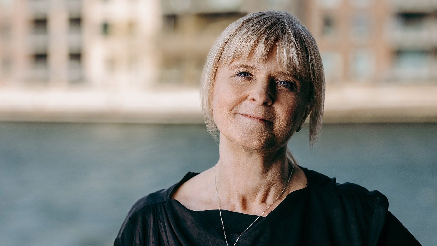 Kvinde med blond hår, smilende udendørs; bygninger og vand i baggrunden.