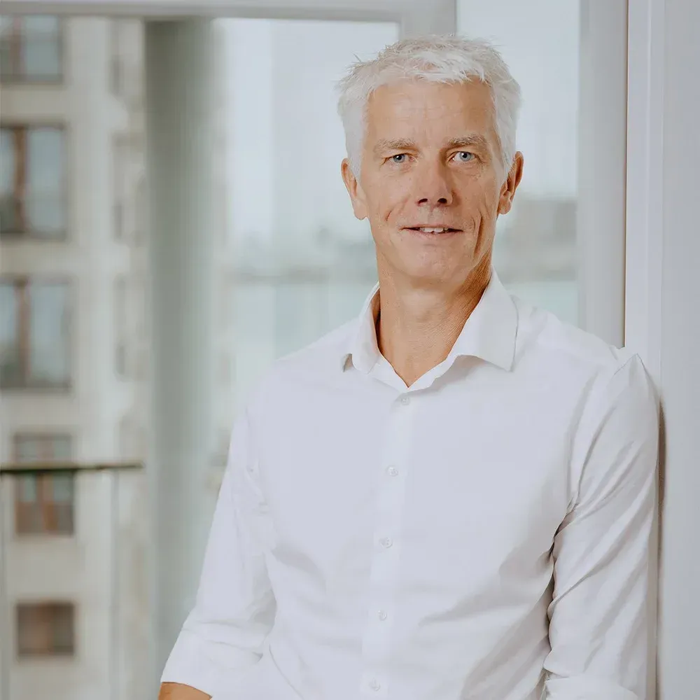 En mand med gråt hår iført en hvid skjorte læner sig op ad en væg og smiler nær et vindue.