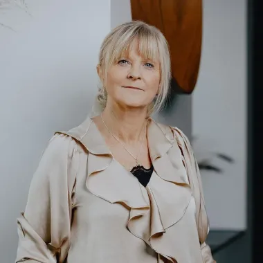 Kvinde i en cremefarvet top, stående indenfor og kigger ind i kameraet.