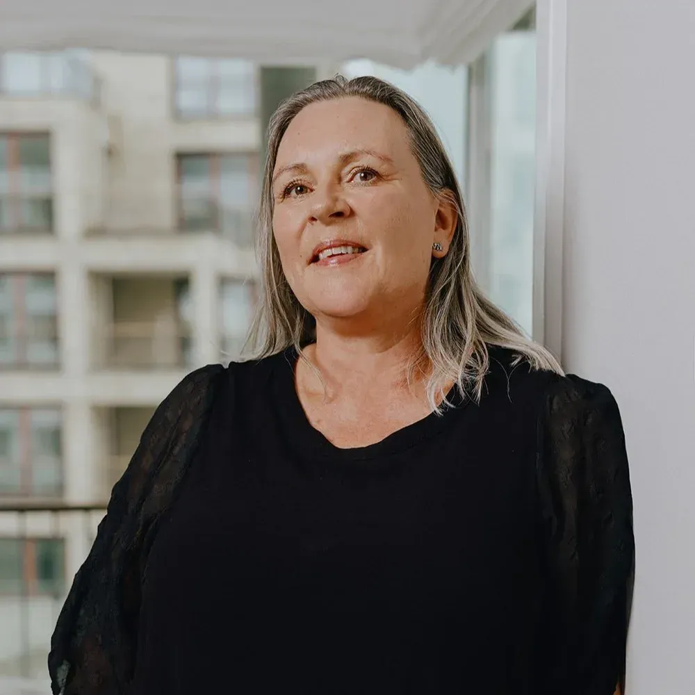 Kvinde med langt gråt hår smiler, iført en sort top, står ved et vindue med udsigt over byen.