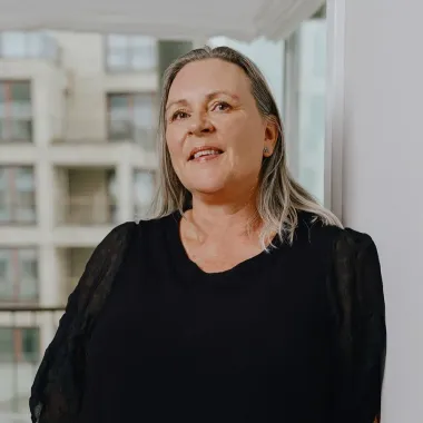 Kvinde med langt gråt hår smiler, iført en sort top, står ved et vindue med udsigt over byen.