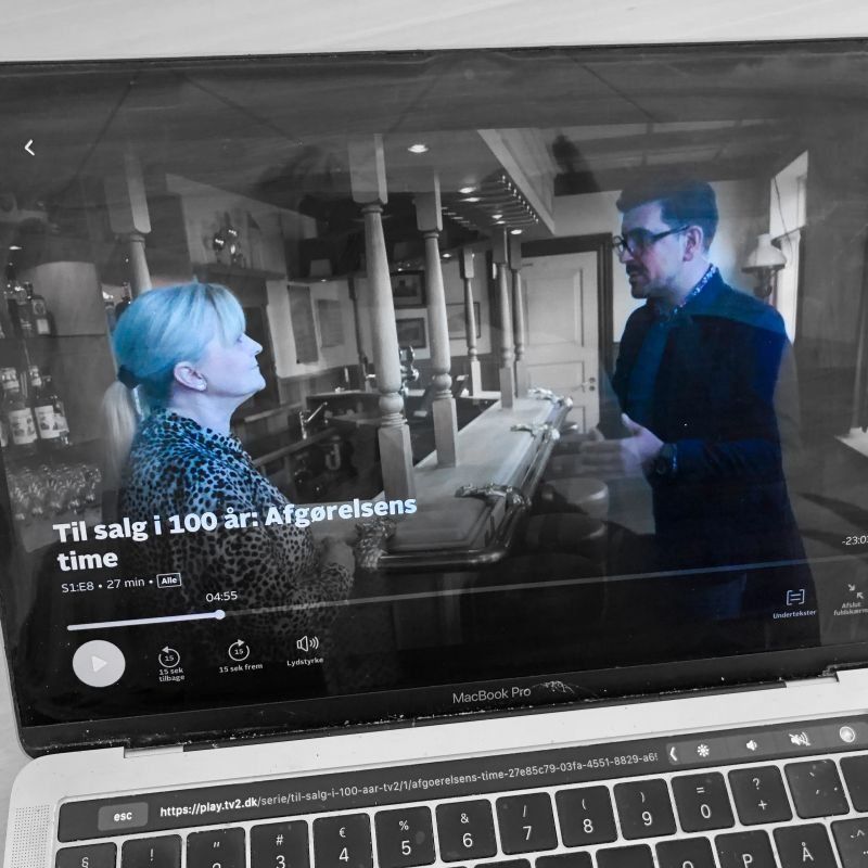 En bærbar computerskærm viser et tv-program med to personer i et køkken. Programmets titel er synlig.