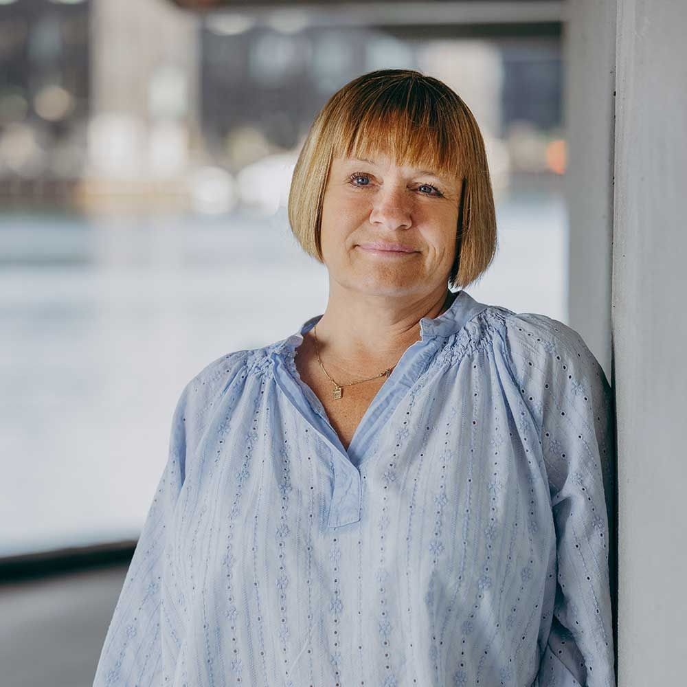 Kvinde med blond bob og pandehår, iført en blå bluse, læner sig op ad en væg udendørs.