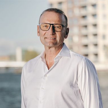 Mand i hvid skjorte og briller smiler, udendørs med bygning og vand i baggrunden.