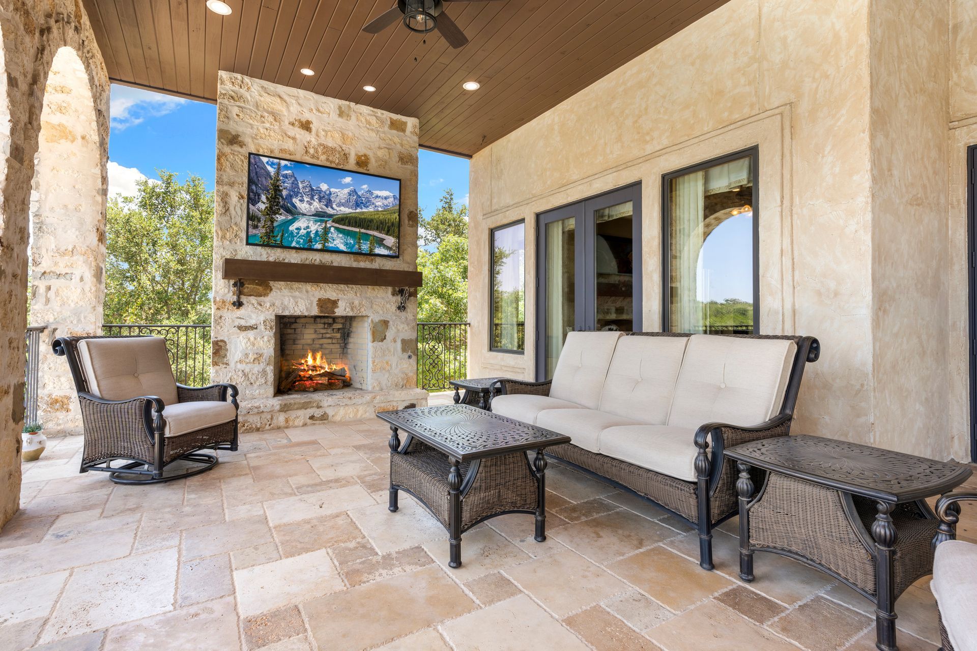 Covered stone patio with a fireplace, television, outdoor sofa, armchair, and coffee tables on a travertine floor.