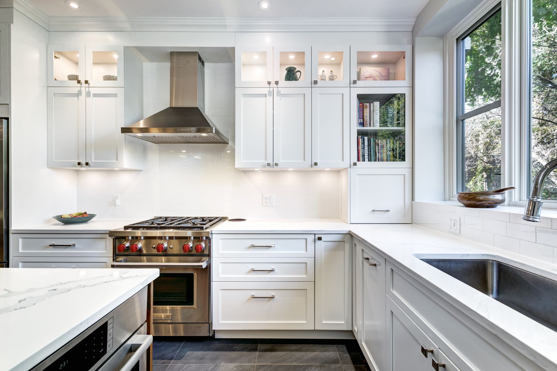 A bright, modern kitchen features white shaker cabinets, a stainless steel range hood, stone countertops, and a sink.