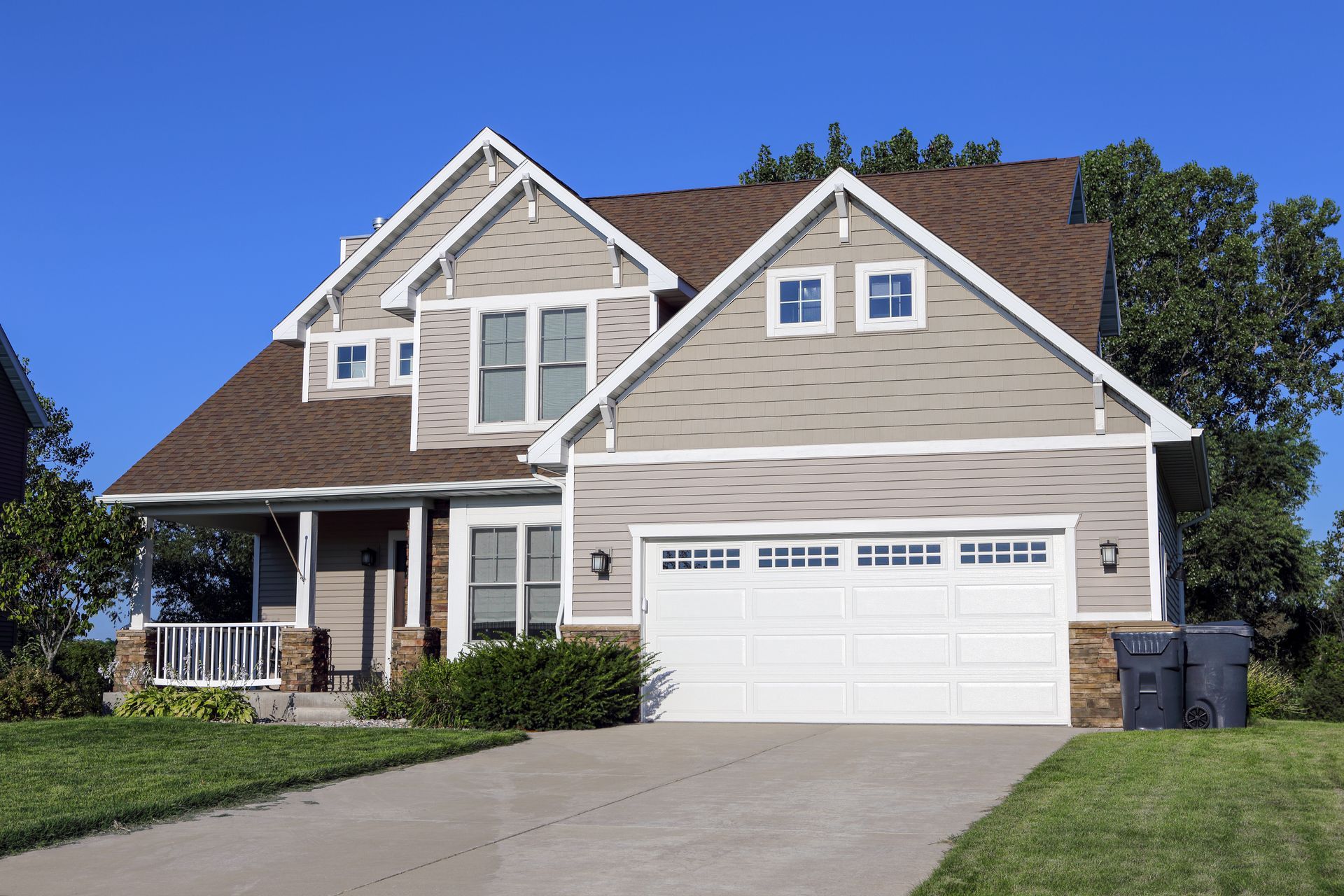A two-story suburban house with light gray siding, brown shingles, and a white two-car garage on a clear, sunny day.