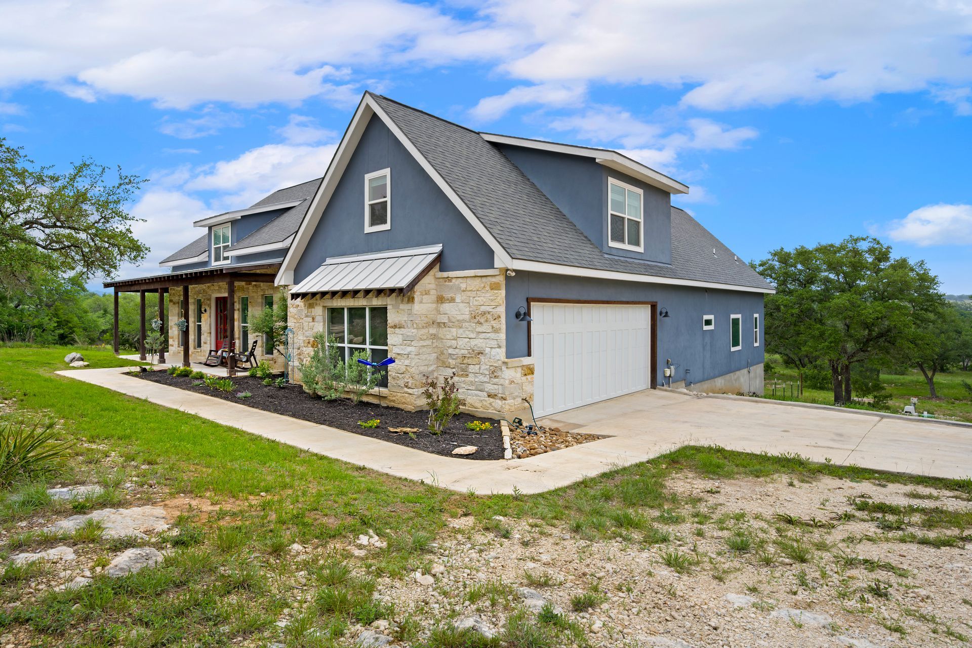 A two-story blue house with stone siding, a white garage, and a wrap-around porch set in a grassy landscape.