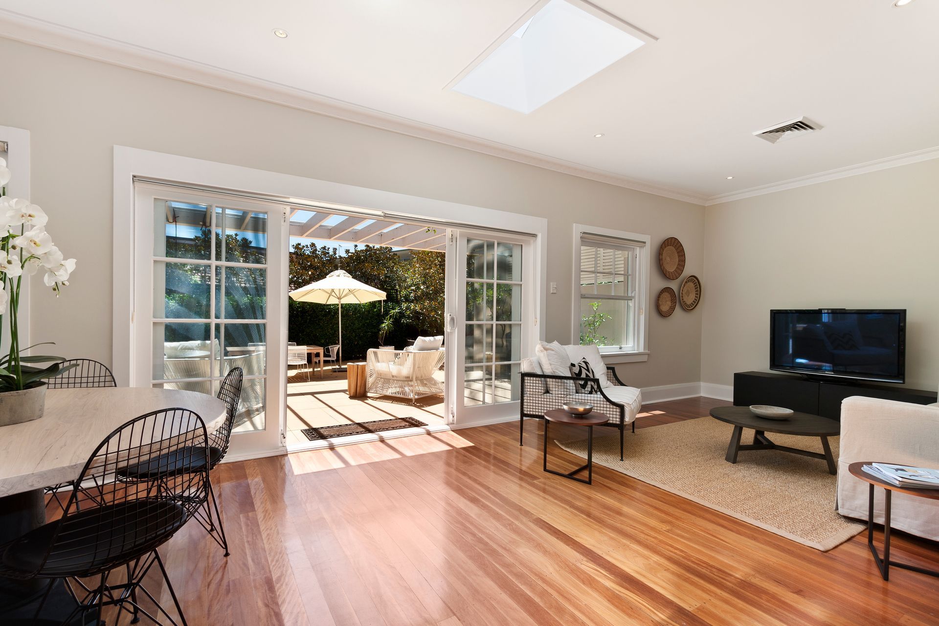 A sunlit living room with wood floors, a dining area, and large glass doors opening onto a patio with outdoor seating.