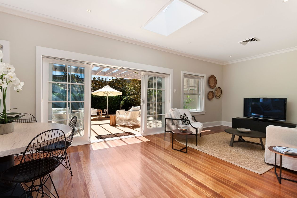 A sunlit living room with wood floors, a dining area, and large glass doors opening onto a patio with outdoor seating.