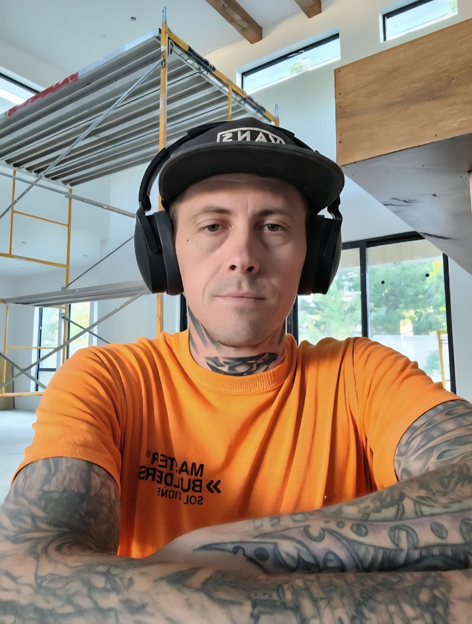 Man in orange shirt and cap, construction site background, arms crossed, tattoos visible.