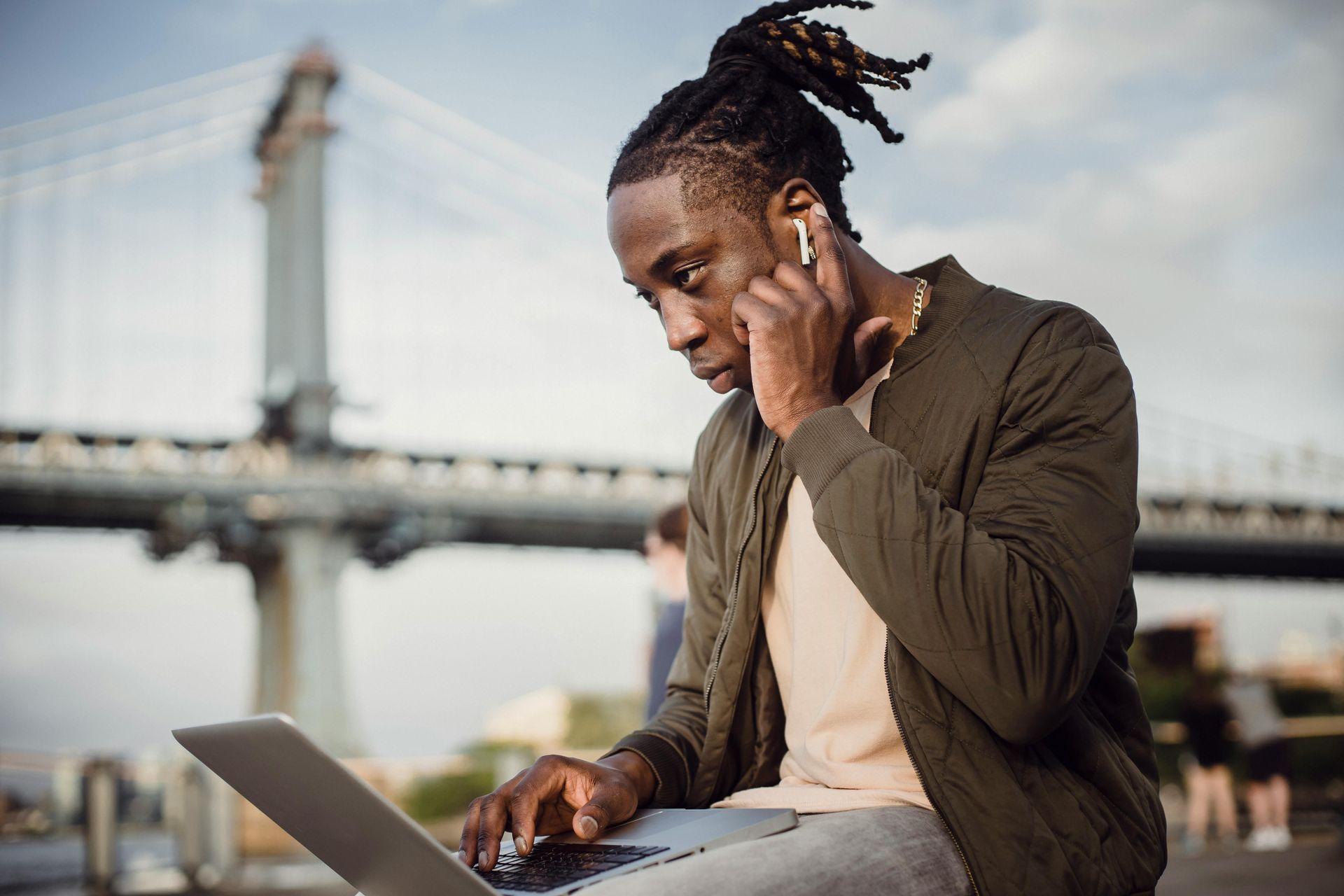 Black man with dreadlocks using laptop, wearing earbuds, near bridge.