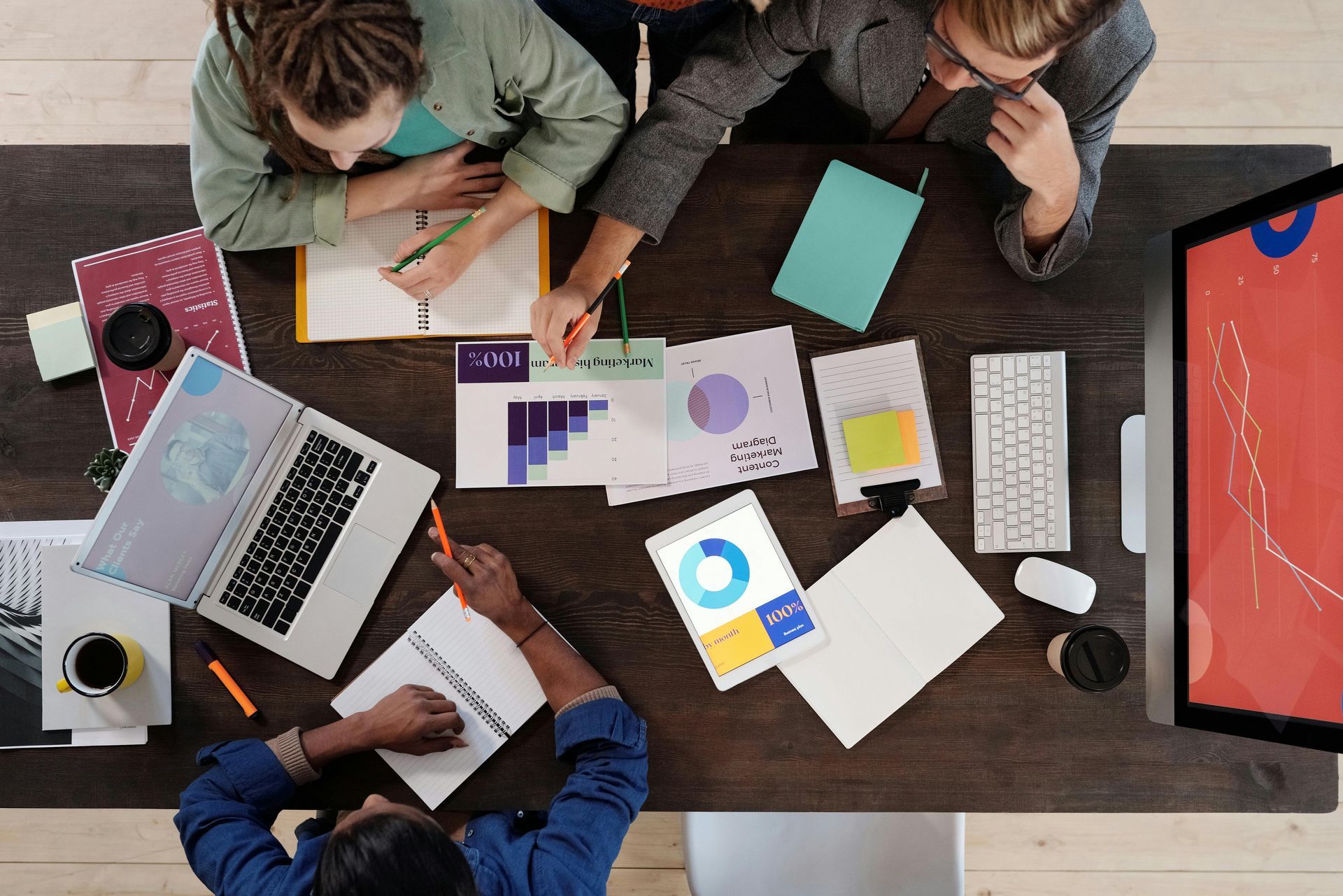 People collaborate at a wooden table with laptops, charts, and notebooks, discussing data in an office.