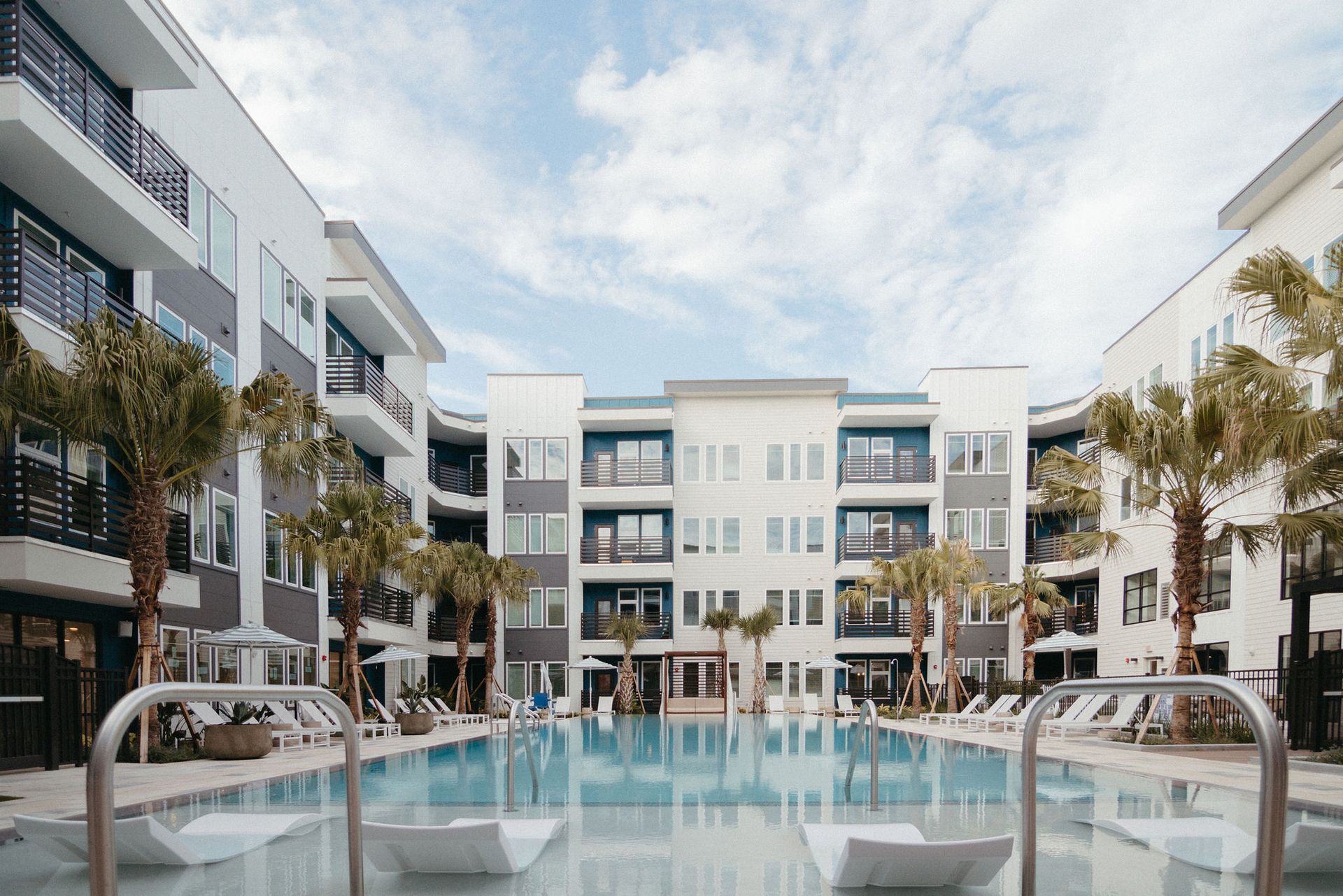 Pool with white lounge chairs between two multi-story apartment buildings on a sunny day.