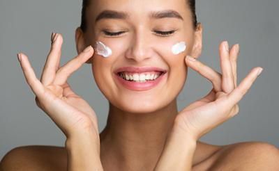 A woman is smiling while applying cream to her face.