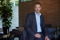 Holger Heid sitzt stehend an der Rücklehne einer Coach im Wohnzimmer und schaut lächelnd direkt in die Kamera