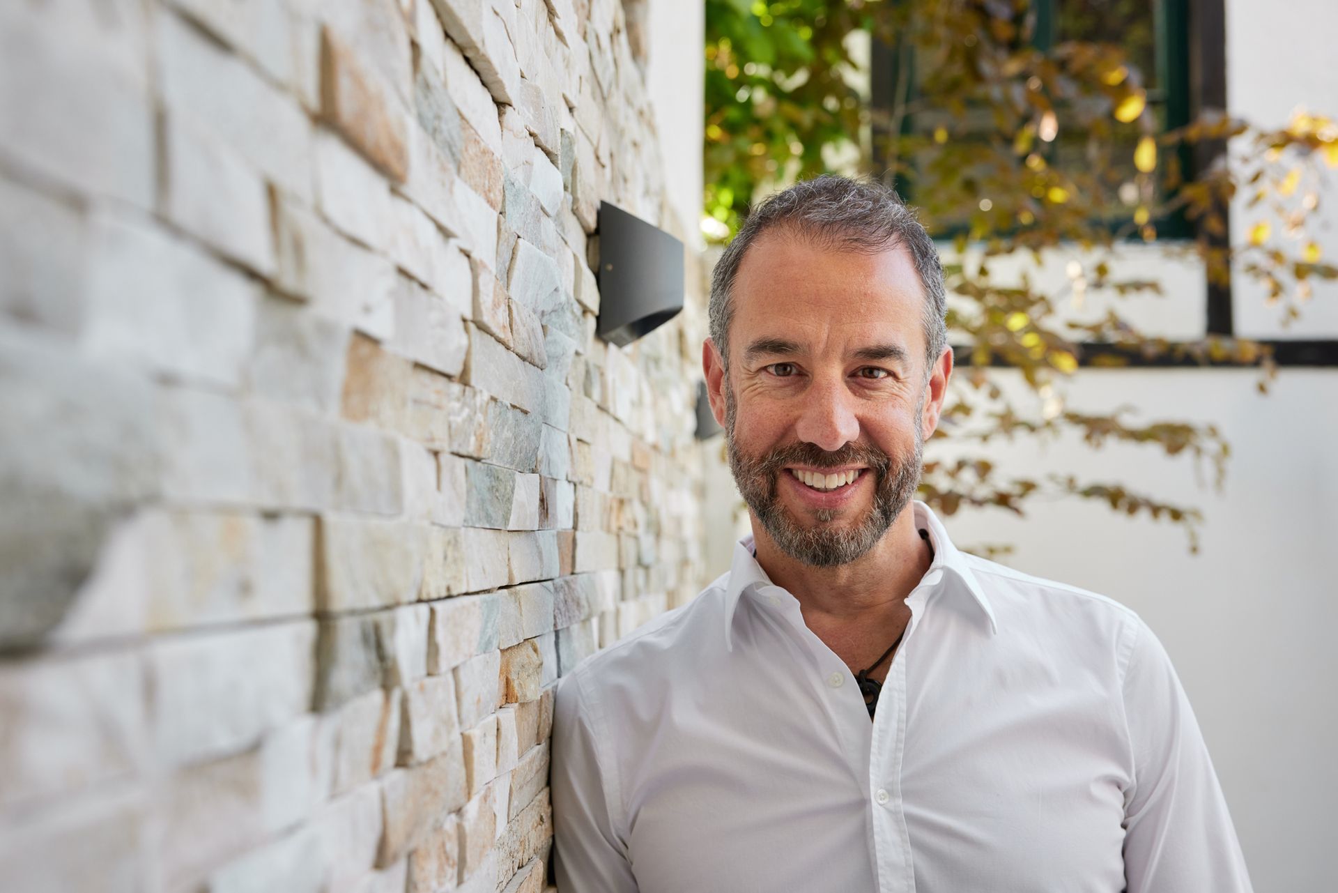 Mentalcoach mit Herz Holger Heid lehnt entspannt mit der rechten Schulter an einer Natursteinwand, lächelt in die Kamera und strahlt Sympathie und Souveränität aus.