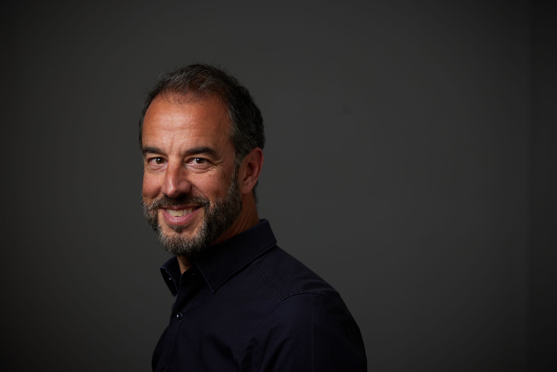 Coach Holger Heid vor einer grauen Wand, Kopf nach links gedreht und in die Kamera schauend, er lächelt