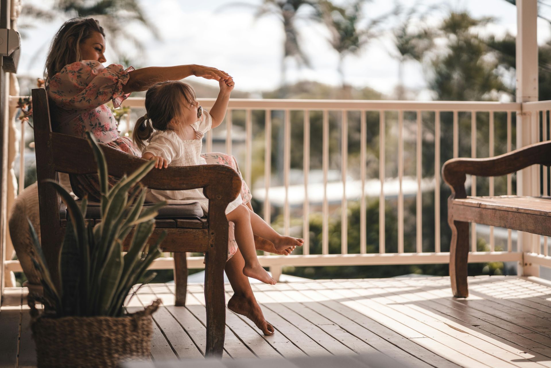 Une mère élégamment vêtue est assise avec sa fille de trois ans, qui a une tétine dans la bouche, sur un banc en bois à l’ombre d’une véranda. C’est l’été et, en arrière-plan, on distingue des palmiers flous.