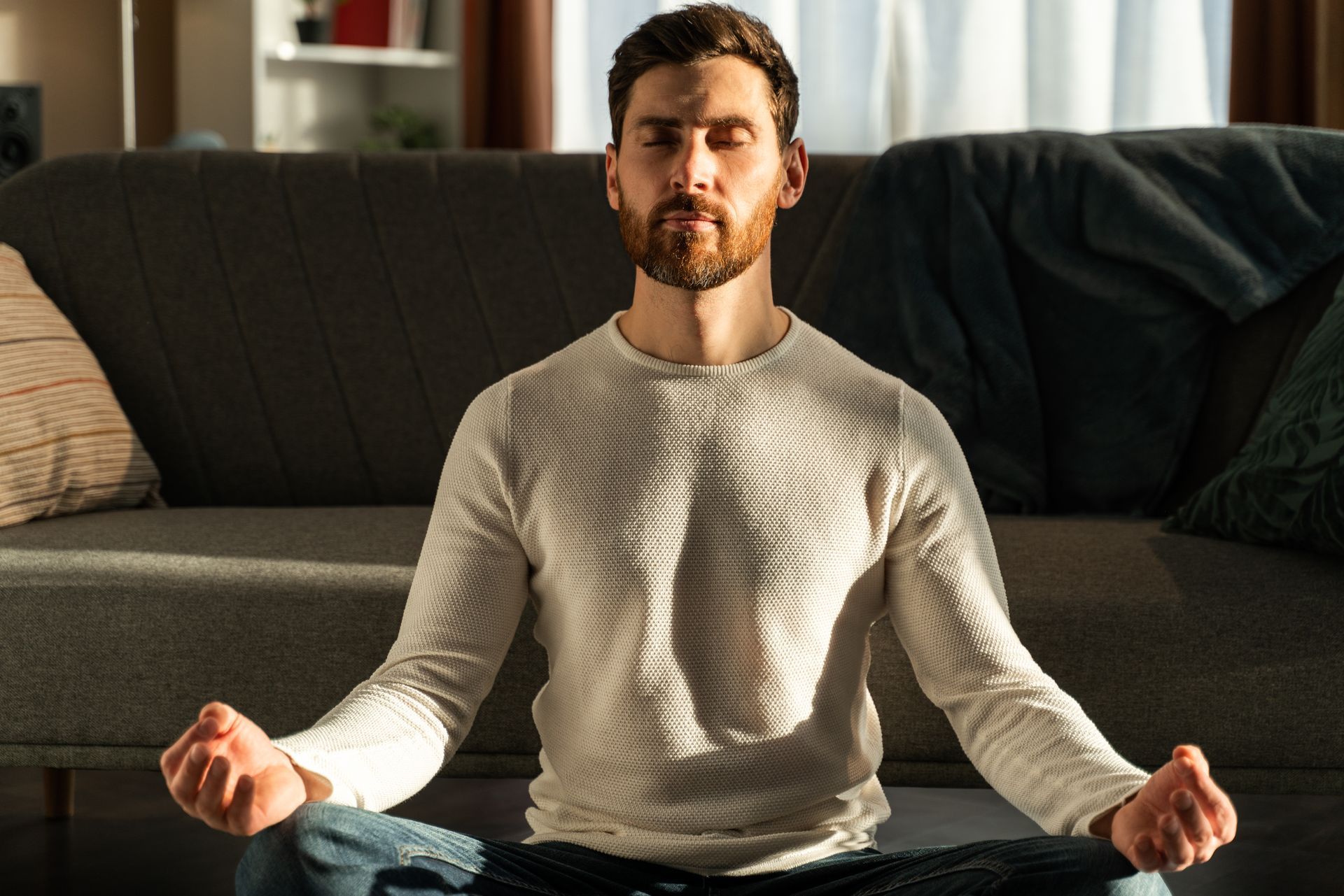 Mentale, innere Stärke Mann meditiert im Schneidersitz auf dem Boden vor seinem Sofa im Wohnzimmer und wird von der Sonne angestrahlt