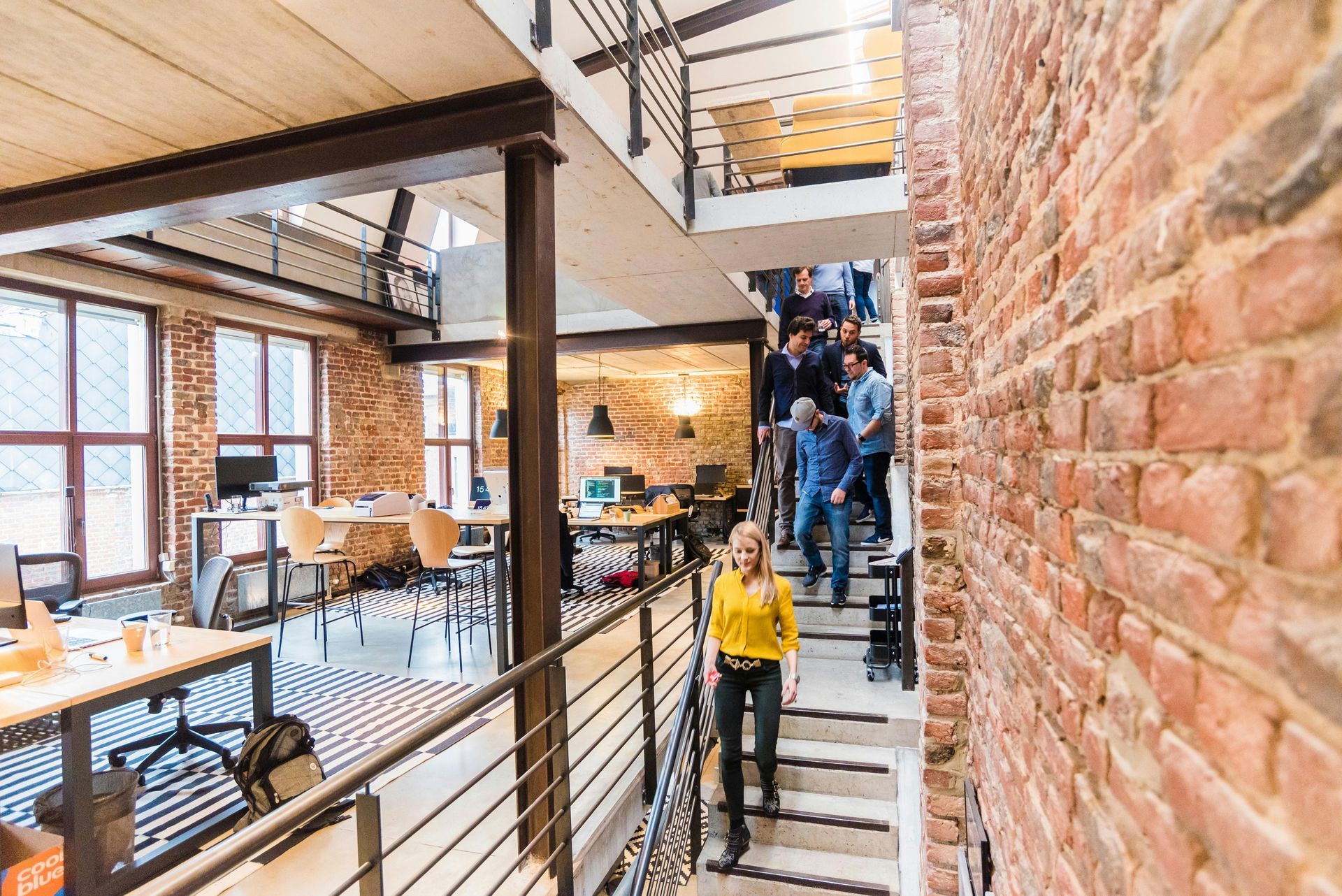 On voit un bureau moderne style industrielle avec des poudres métalliques et des briques. Un groupe d'employé descend l'escalier.
