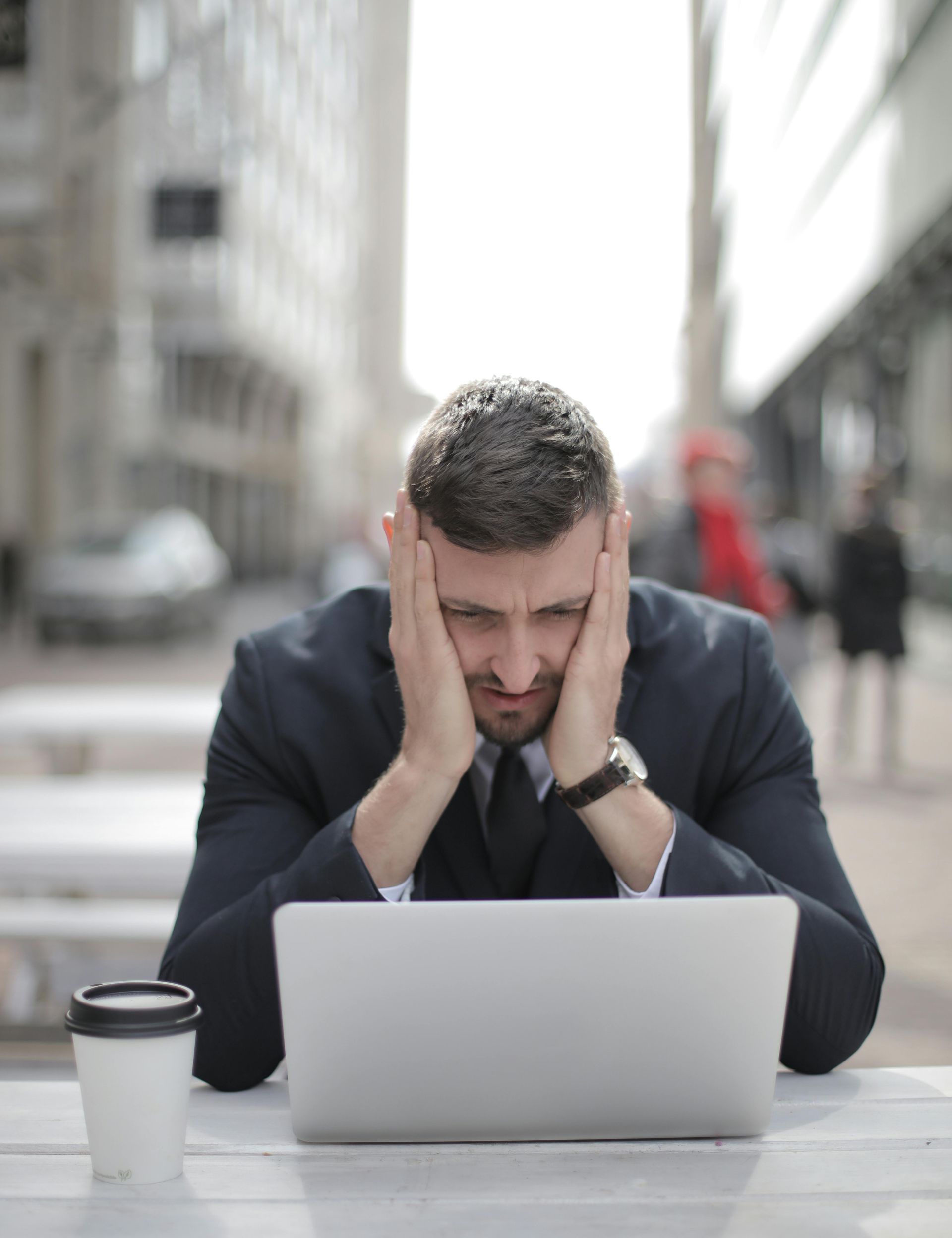 Müdigkeit, Stress, Überlastung, Burnout: ein schleichender Prozess Ein Mann Mitte 30 sitzt im Anzug an einem Tisch im Freien, stützt sein Gesicht in beide Hände und wirkt müde und erschöpft, überfordert und hilflos. Er schaut leer in das aufgeklappte Laptop vor ihm.