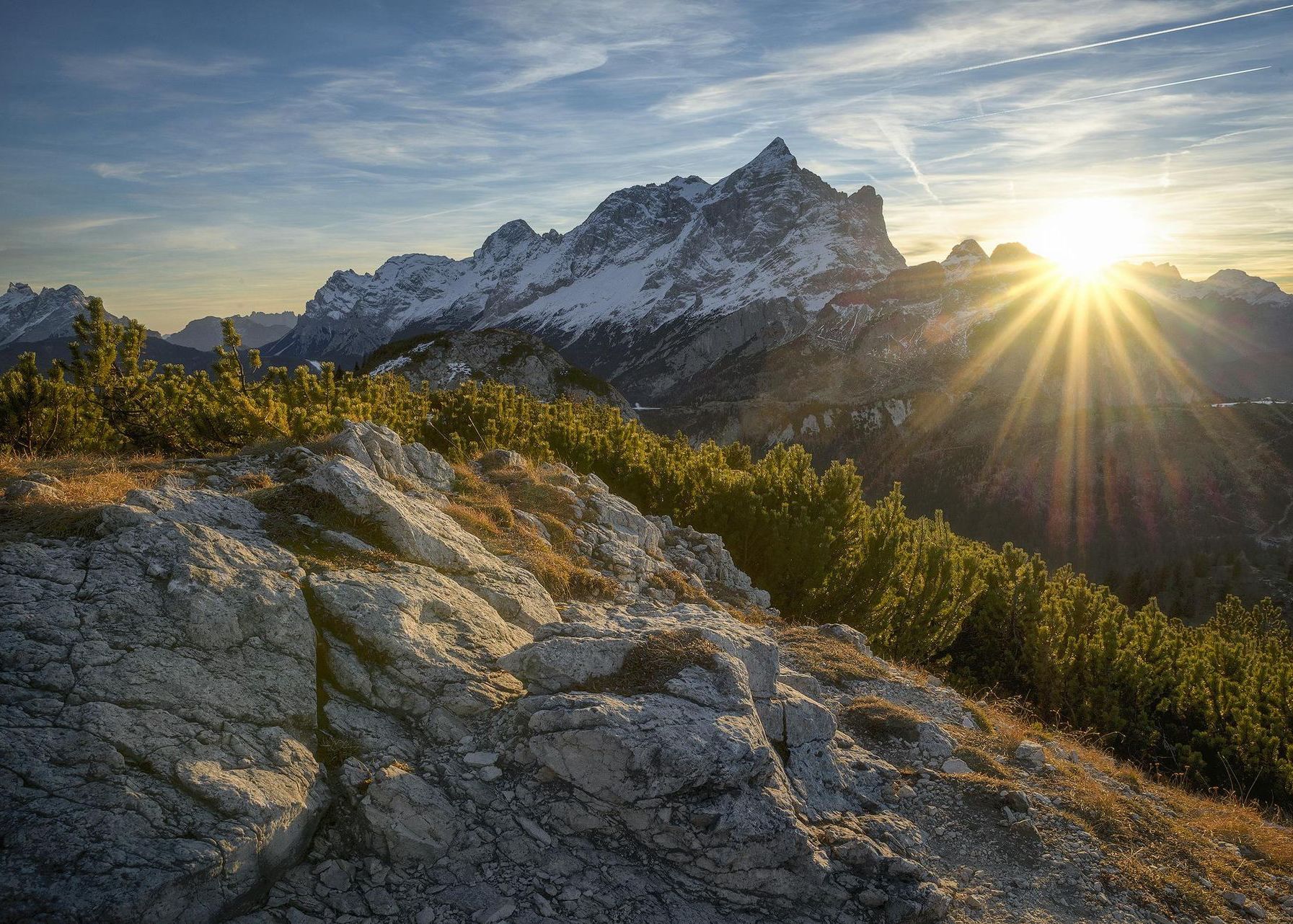 Natur und körperliche Betätigung als Ausgleich Die Sonne geht gerade hinter einer Bergkette unter und spendet noch warme Strahlen auf den Berg, wo sich der Fotograf befindet. Es scheint ein schöner Frühsommertag zu sein, nur wenige Schleierwolken sind am Himmel. Auf der Schattenseite der Bergkette liegt noch Schnee.