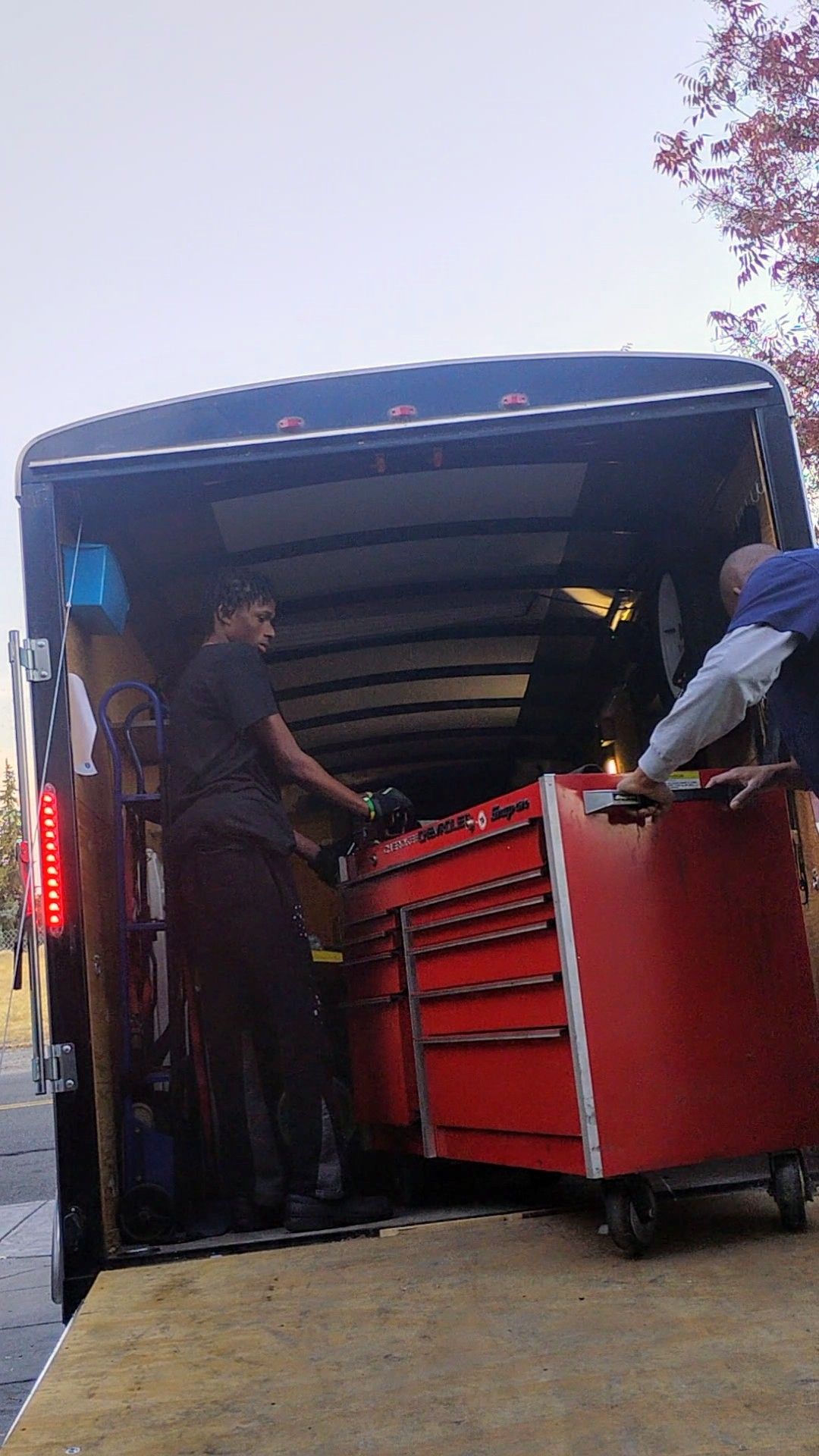 Two men are loading a red toolbox into a trailer.