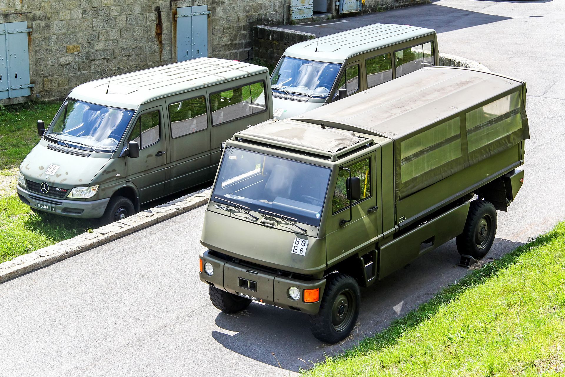 Three olive green military vehicles on a paved road: a truck with a flatbed and two vans.
