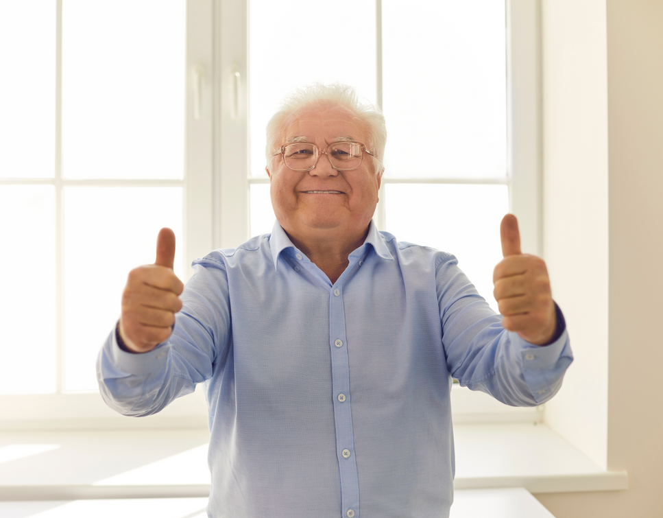 An elderly man is giving two thumbs up in front of a window.