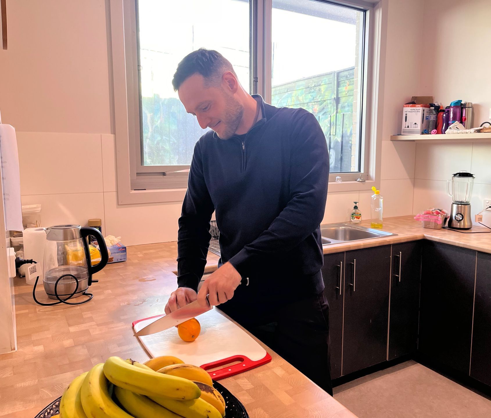 A man is cutting an orange on a cutting board in a kitchen.