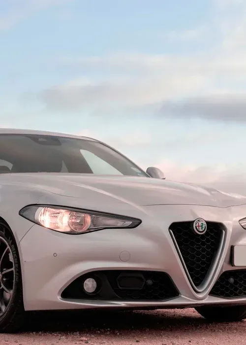 A low-angle view of a silver Alfa Romeo sedan parked on a gravel surface under a soft, cloudy sky.