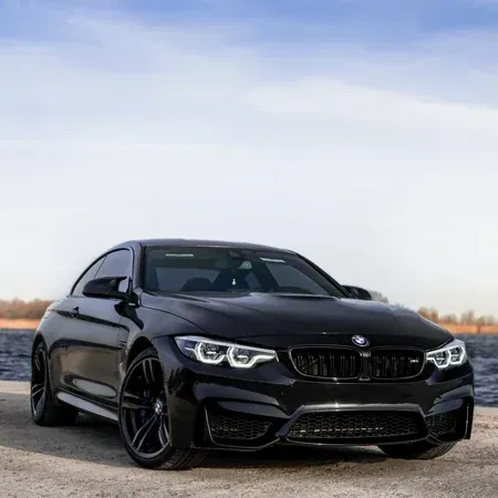 A black BMW M4 coupe parked on a gravel surface near a body of water under a light blue sky.