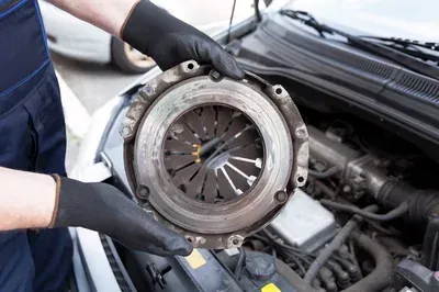 A technician in blue coveralls works on the engine of a grey car with its hood open inside an auto repair shop.