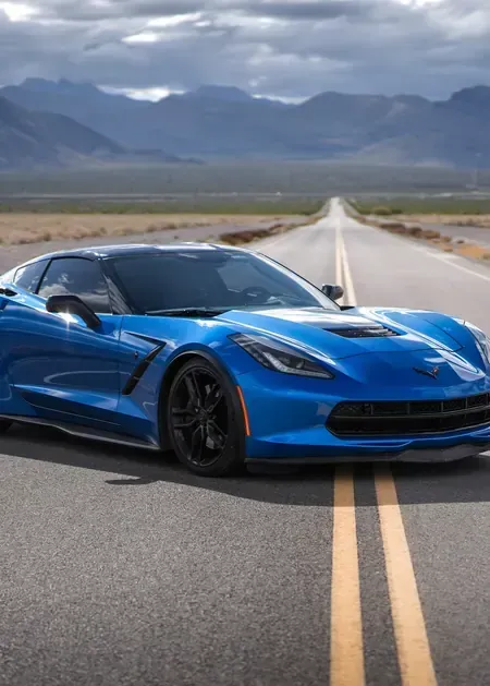 A blue Chevrolet Corvette parked on a long, empty road stretching toward distant mountains under a cloudy sky.