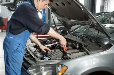 A technician in blue coveralls works on the engine of a grey car with its hood open inside an auto repair shop.