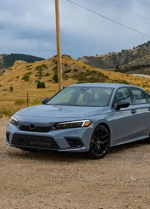A grey Honda Civic sedan parked on a gravel road against a backdrop of rolling, arid hills under a cloudy sky.