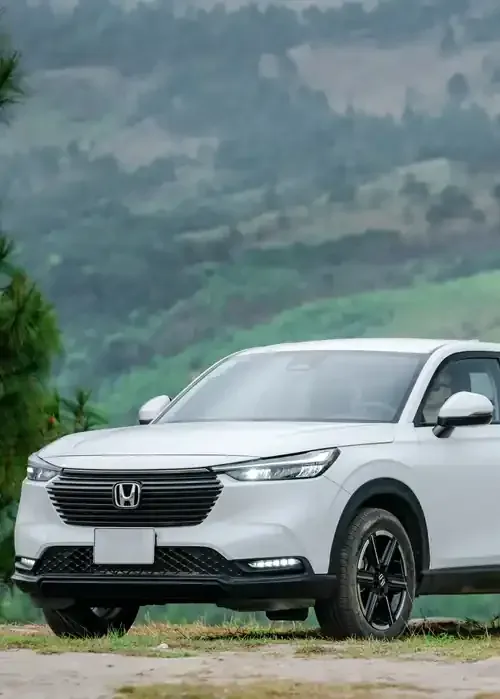 A white Honda HR-V SUV parked on a grassy hill with a scenic, forested mountain landscape in the background.