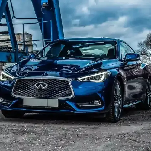 A dark blue Infiniti coupe parked on a gravel surface beneath a blue industrial crane structure under a cloudy sky.